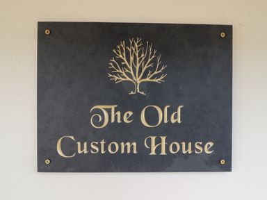 A black sign with gold lettering and a tree design that reads The Old Custom House at The Old Custom House in Wangford