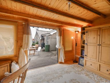 A wooden room with a large cabinet a floor lamp and an open door leading to a patio with chairs at The Old Custom House in Wangford