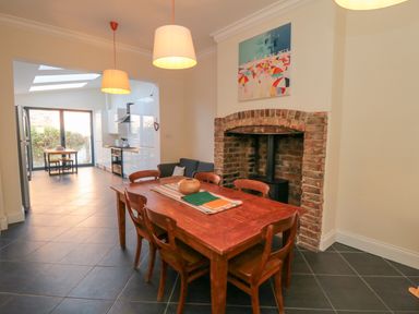 A dining area with a wooden table and chairs next to a brick fireplace opening and a kitchen area with white cabinets and skylights in the background at South Bay House in Scarborough