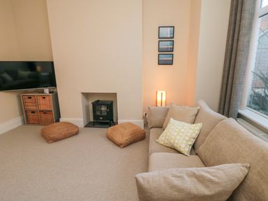 A living room with a beige sofa and cushions a small black stove two woven floor cushions a lamp and a wall-mounted TV at South Bay House in Scarborough