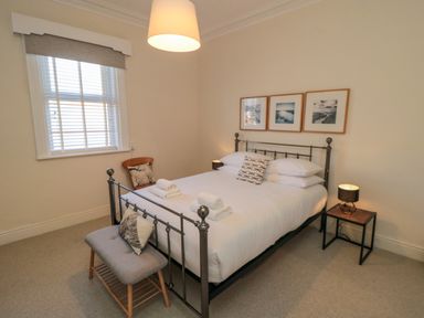 A bedroom with a metal bed frame a bench with cushions a chair a window with blinds and framed pictures on the wall at South Bay House in Scarborough