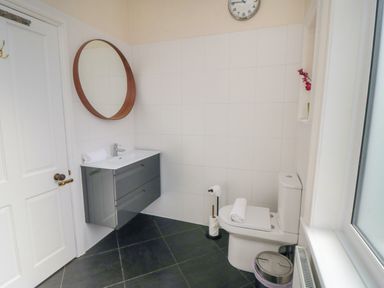 A bathroom with a wall-mounted sink, round mirror, toilet, small trash can, and clock at South Bay House in Scarborough