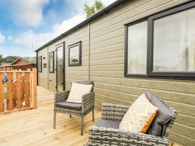 An outdoor wooden deck with two wicker chairs with cushions next to a green building with black windows at Ashton Lodge - Sherwood 4 in South Lakeland Leisure Village