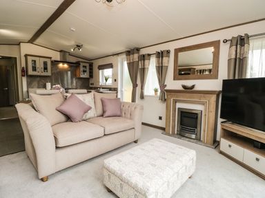 A living room with a beige sofa purple cushions an ottoman a fireplace a flat screen tv and kitchen cabinets at Ashton Lodge - Sherwood 4 South Lakeland Leisure Village