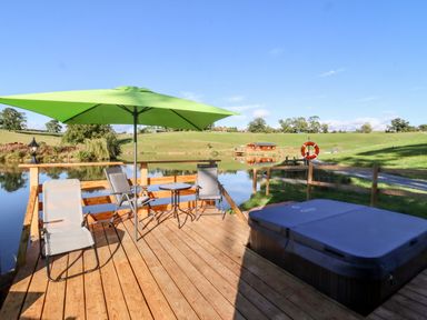 A deck with chairs table and a hot tub next to a lake with green hills at Goose Pod Near Clifton Upon Teme