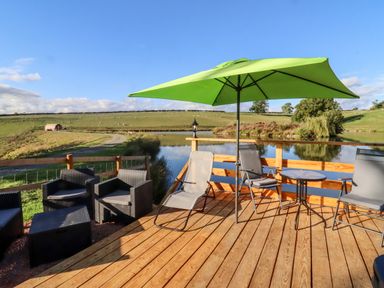 An outdoor wooden deck with chairs a small table and a green umbrella by a pond at Goose Pod Near Clifton Upon Teme