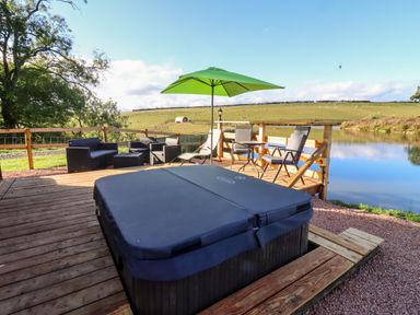 An outdoor deck with a covered hot tub chairs tables and a green umbrella by a pond at Goose Pod Near Clifton Upon Teme
