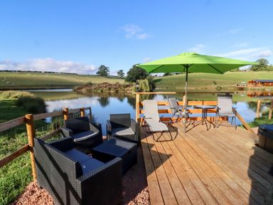 An outdoor deck with seating including black wicker chairs and a green umbrella near a lake at Goose Pod Near Clifton Upon Teme