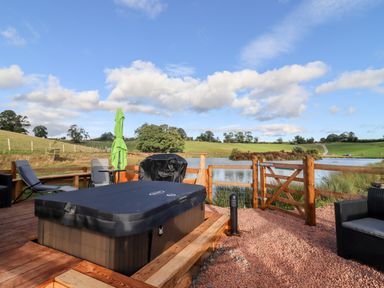 An outdoor wooden deck with a covered hot tub chairs a green umbrella and a wooden fence overlooking a pond at Gander Pod Near Clifton Upon Teme