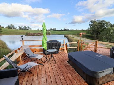 A wooden deck with lounge chairs a small table a green umbrella a covered barbecue and a hot tub by a pond at Gander Pod Near Clifton Upon Teme