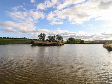 A lake with small islands trees and grasslands under a partly cloudy sky at Gander Pod Near Clifton Upon Teme