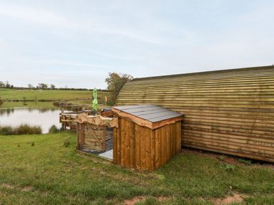 A wooden pod structure near a small pond with grassy fields in the background at Gander Pod Near Clifton Upon Teme