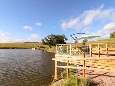 A wooden deck with a picnic table and umbrella by a lake with grassy fields and trees at Cedar Cabin Near Clifton Upon Teme