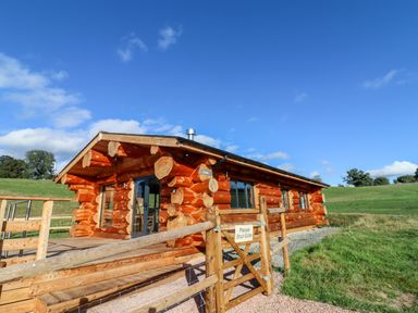 A wooden log cabin with a deck and a gate in a grassy landscape at Cedar Cabin Near Clifton Upon Teme