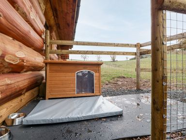An outdoor enclosed area with a wooden dog house a gray mat two metal bowls and a wooden log building at Cedar Cabin Near Clifton Upon Teme