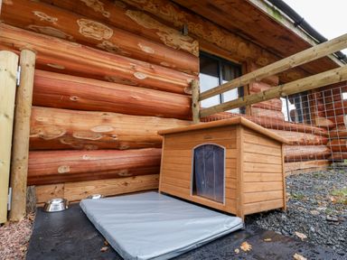 A wooden dog house with a mat and metal bowls outside a log cabin at Cedar Cabin Near Clifton Upon Teme