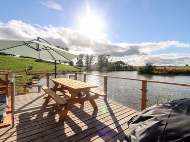 A wooden picnic table with a large white umbrella on a deck overlooking a lake with trees and grassy hills near Douglas Cabin Near Clifton Upon Teme