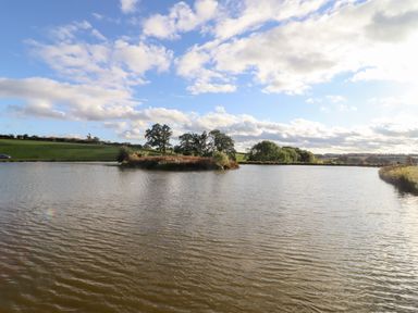 A lake with small islands and trees surrounded by grassy fields under a partly cloudy sky at Douglas Cabin Near Clifton Upon Teme