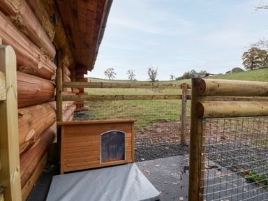 A small fenced outdoor area with a wooden dog house and a mat beside a log cabin at Douglas Cabin Near Clifton Upon Teme