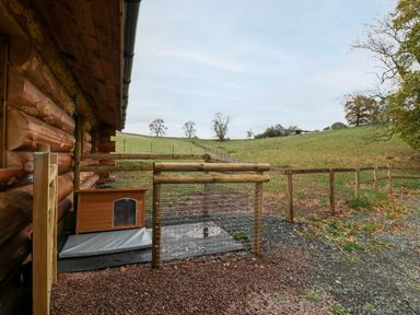 A fenced outdoor area next to a log cabin with a small wooden shelter and a rural field beyond at Douglas Cabin Near Clifton Upon Teme