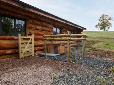 An outdoor area with a wooden cabin and a fenced enclosure containing a dog house near a field with a tree at Douglas Cabin Near Clifton Upon Teme