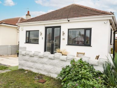 A small white house with black window frames and doors behind a low stone wall with plants in front at Cove House in Kinmel Bay