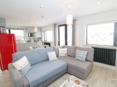 An open plan living room and kitchen with a grey sofa, round dining table, red refrigerator and large windows at Cove House in Kinmel Bay