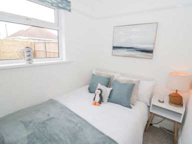 A bedroom with a bed with pillows and a stuffed penguin a bedside table with a lamp and a window with a ceramic pitcher on the sill at Cove House in Kinmel Bay