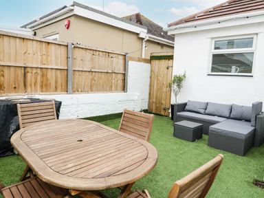 An outdoor patio area with a wooden table and chairs a gray cushioned sofa and small tables on artificial grass at Cove House in Kinmel Bay