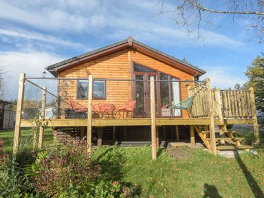 A wooden cabin with a raised deck and chairs in a grassy area at Oaks One Swarland near Felton