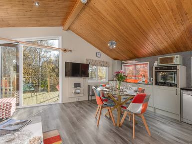 A kitchen and dining area with a glass table set for four and a wall-mounted TV with a door opening to an outdoor deck at Oaks One Swarland near Felton