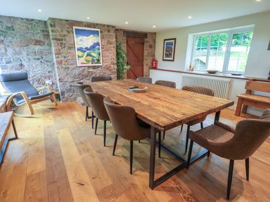 A dining room with a large wooden table surrounded by brown chairs a rocking chair against a stone wall and windows at Flaska House in Troutbeck Near Penrith