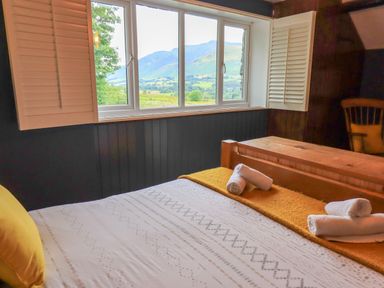 A bedroom with a bed with white and yellow bedding a wooden chair and a window with shutters showing a mountain view at Flaska House in Troutbeck Near Penrith