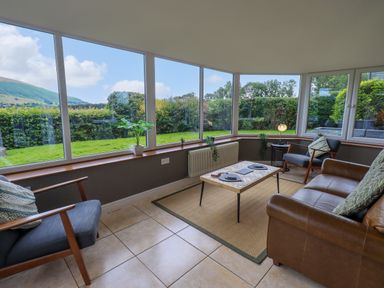 A seating area with chairs a sofa and a coffee table by large windows overlooking a garden at Flaska House in Troutbeck Near Penrith