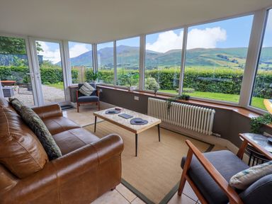 A sunroom with a brown leather couch two gray chairs a coffee table and large windows showing mountains at Flaska House in Troutbeck Near Penrith