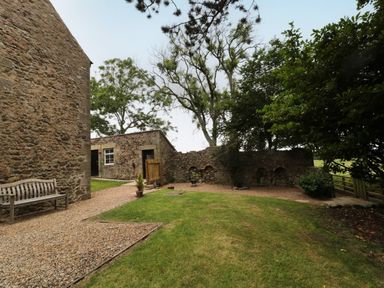 An outdoor area with stone buildings a wooden bench gravel paths grass and trees at 1 Roddam Rigg Cottage in Roddam near Wooler