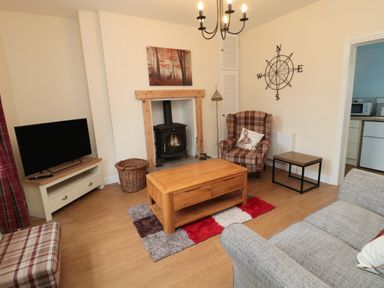 A living room with a wooden coffee table a plaid armchair a gray sofa a wood stove and a television at 1 Roddam Rigg Cottage in Roddam near Wooler
