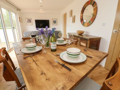 A wooden dining table set with plates bowls wine glasses and a wine bottle in a room with a colorful round mirror a wooden cabinet and a wall-mounted TV at Syrah Seaview