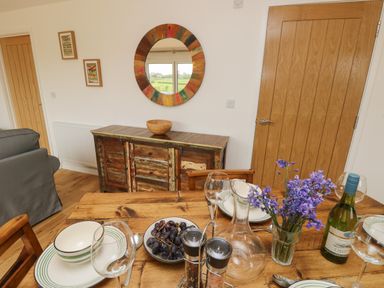 A dining table set with plates bowls wine glasses a bowl of grapes a vase with purple flowers a bottle of wine and a wooden sideboard with a round mirror on the wall at Syrah Seaview