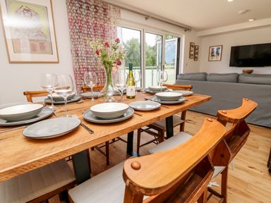 A dining table set with plates glasses and a bottle of wine in a room with wooden chairs a sofa and a wall mounted television at Syrah Seaview
