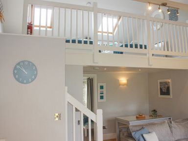 A living area with a gray couch, a dining table with chairs, wall clock, and a loft with beds at 5 Keeper's Cottage, Hillfield in Dartmouth