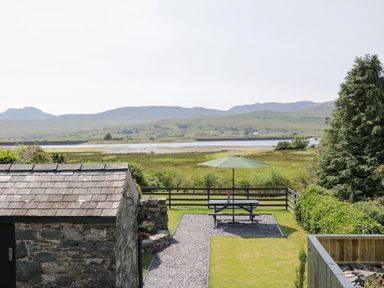 A garden with a picnic table and green umbrella near a stone building and overlooking a grassy field with hills beyond at Minffordd in Trawsfynydd
