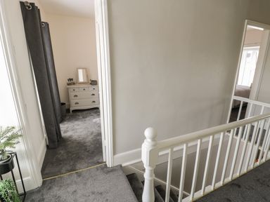 A hallway with a staircase railing leading to two rooms one with a gray chest of drawers and curtains at Minffordd in Trawsfynydd