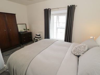 A bedroom with a bed covered in light bedding a window with gray curtains a dark wooden wardrobe a dark wooden dresser with a mirror a white chair with folded towels at Minffordd in Trawsfynydd