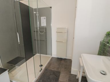 A bathroom with a glass shower, white sink on a stand, towel radiator, and grey floor mat at Minffordd in Trawsfynydd
