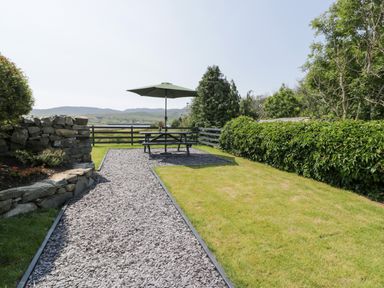 An outdoor garden area with a gravel path leading to a picnic table with an umbrella and green foliage around at Minffordd in Trawsfynydd