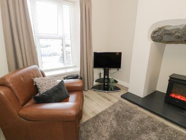 A living area with a brown leather armchair, pillows, a TV on a stand, a window with blinds and curtains, and a fireplace with an electric stove at Minffordd in Trawsfynydd