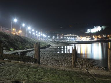 A nighttime view of a beach with streetlights along a promenade and a brightly lit castle in the background at The Headlands - Gower in The Mumbles