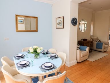 A dining area with a round table set with plates and glasses and cream chairs next to a living area with armchairs and a wooden sideboard at The Headlands - Gower in The Mumbles