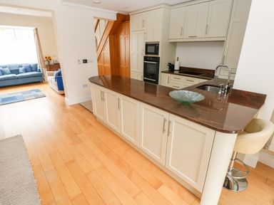 A kitchen with brown countertops white cabinets and a beige bar stool next to a living room with blue sofas at The Headlands - Gower in The Mumbles